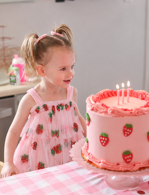 Girl wearing a pink strawberry sequin tulle dress beside a strawberry birthday cake and pink gingham tablecloth